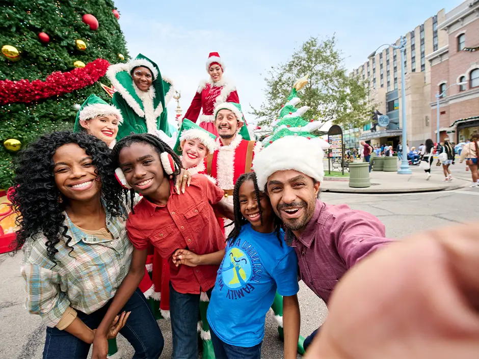 a Smiling Family Takes a Selfie in Front of a Large Christmas Tree at Universal Studios Orlando, Joined by Festive Performers in Elf and Santa Costumes on a Sunny City Street—capturing the Magic of the Holidays with People and Buildings in the Background. - Favorite Grampy Travels - Favorite Grampy Travels 2026
