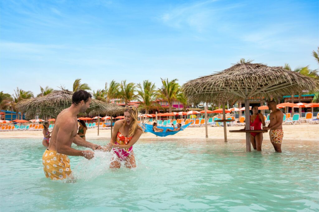 Four adults enjoy a sunny day at a Royal Caribbean beach resort; two play and splash in shallow water while two others chat under a thatched umbrella. Colorful chairs, palm trees, and blue sky complete the lively scene. - Favorite Grampy Travels