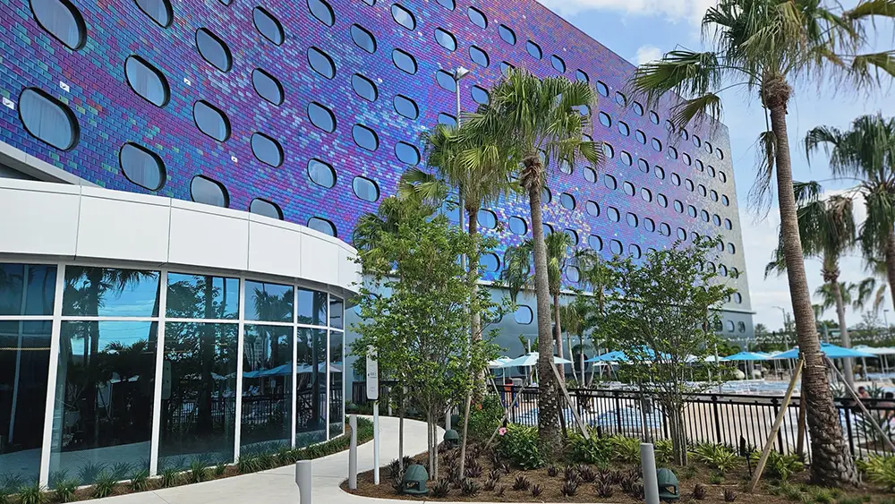 a Modern Building with a Shimmering Purple Facade and Round Windows Stands Behind Palm Trees and a Pool Area, with Clear Blue Sky Overhead and Lounge Chairs Visible Near the Water. - Favorite Grampy Travels - Favorite Grampy Travels 2026