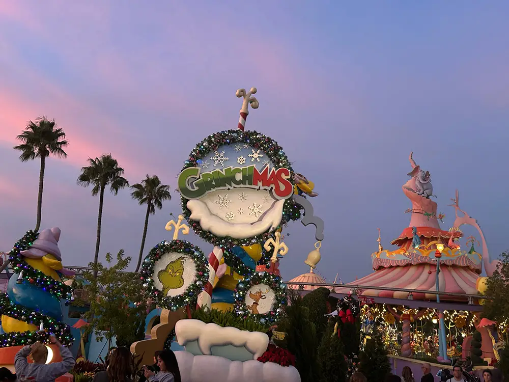 a Whimsical, Colorful Christmas Display with a Grinchmas Sign, Candy Cane Decorations, and Festive Lights, Set Against a Twilight Sky with Palm Trees and Dr. Seuss-inspired Buildings in the Background. - Favorite Grampy Travels - Favorite Grampy Travels 2026
