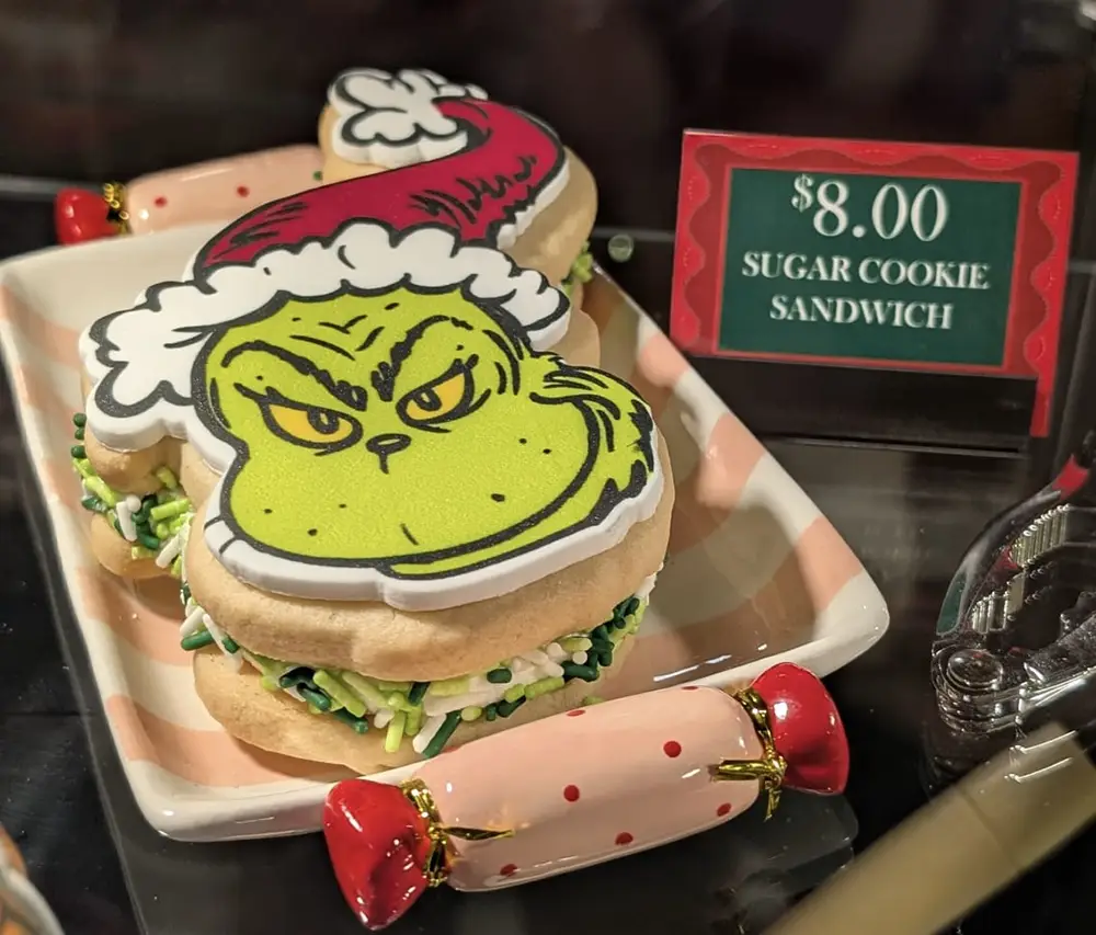 a Festive Sugar Cookie Sandwich Decorated with the Grinchs Face in Icing Sits on a Plate, Filled with Green and White Sprinkles. a Sign Beside It Reads .00 Sugar Cookie Sandwich. - Favorite Grampy Travels - Favorite Grampy Travels 2026