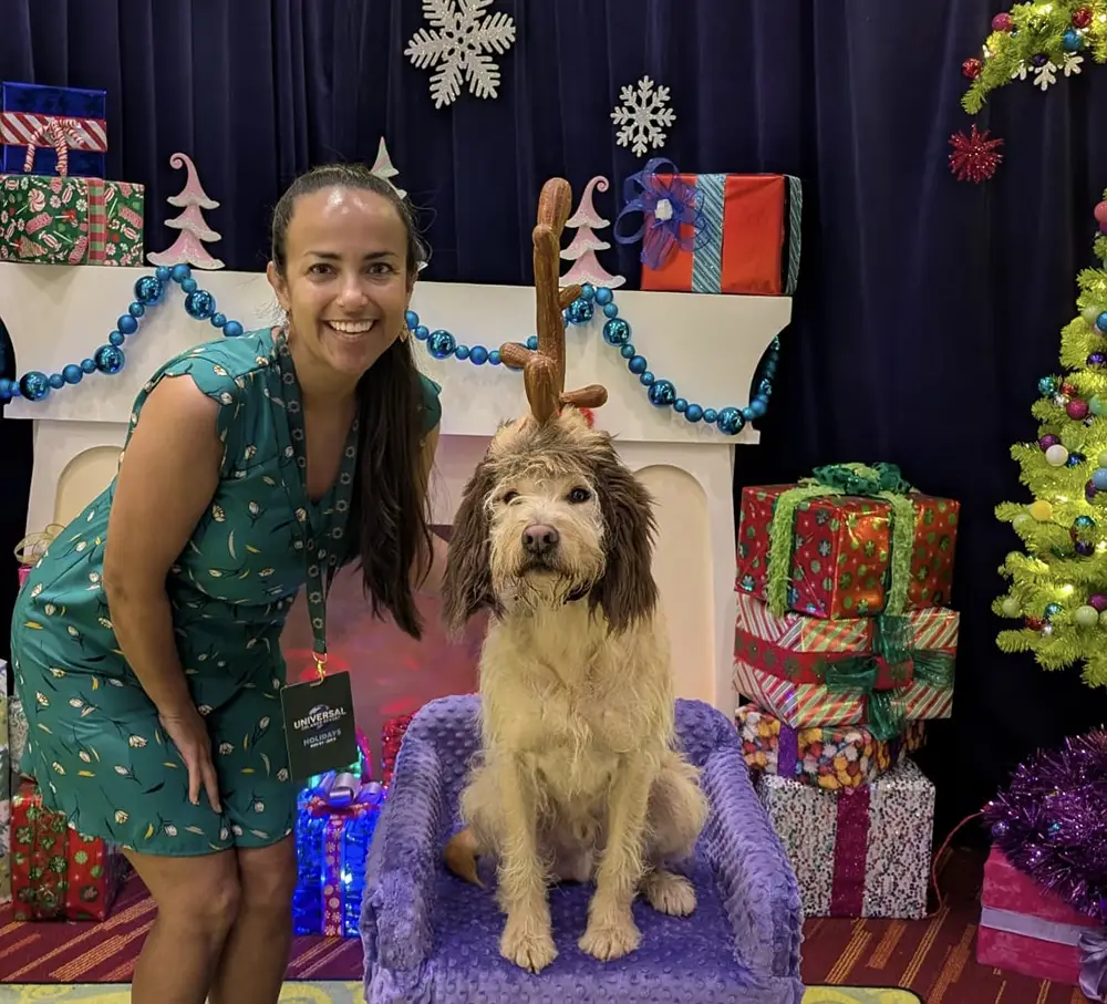 a Woman Kneels Next to a Fluffy Dog Wearing Reindeer Antlers. They Are in Front of a Festive Holiday Backdrop with Wrapped Presents, a Decorated Tree, and Blue Garland. the Woman is Smiling and Wearing a Green Dress. - Favorite Grampy Travels - Favorite Grampy Travels 2026
