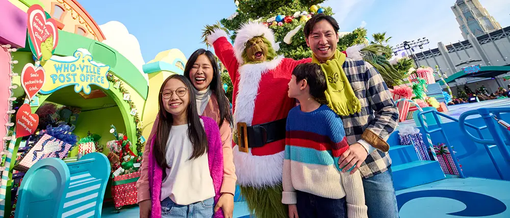 a Family of Four Smiles and Poses with a Person Dressed As the Grinch in a Santa Costume at a Festive, Colorful Outdoor Holiday Event, with Decorations and a Who-ville Post Office in the Background. - Favorite Grampy Travels