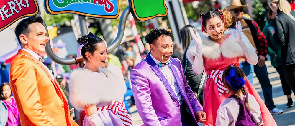 at Universal Studios Hollywood Four Whos Wearing Colorful Suits and Festive Dresses Smile in Whoville and Interact with a Young Child at the Grinchmas Holiday Parade, with Bright “merry Christmas” Signs Overhead. - Favorite Grampy Travels
