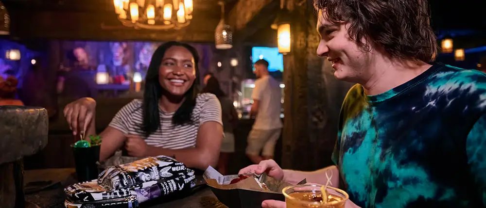 Two People Sit at a Dimly Lit Restaurant Table, Smiling and Enjoying Drinks and Food. the Background Features Warm Lighting, Rustic Decor, and Other Diners. - Favorite Grampy Travels - Favorite Grampy Travels 2026