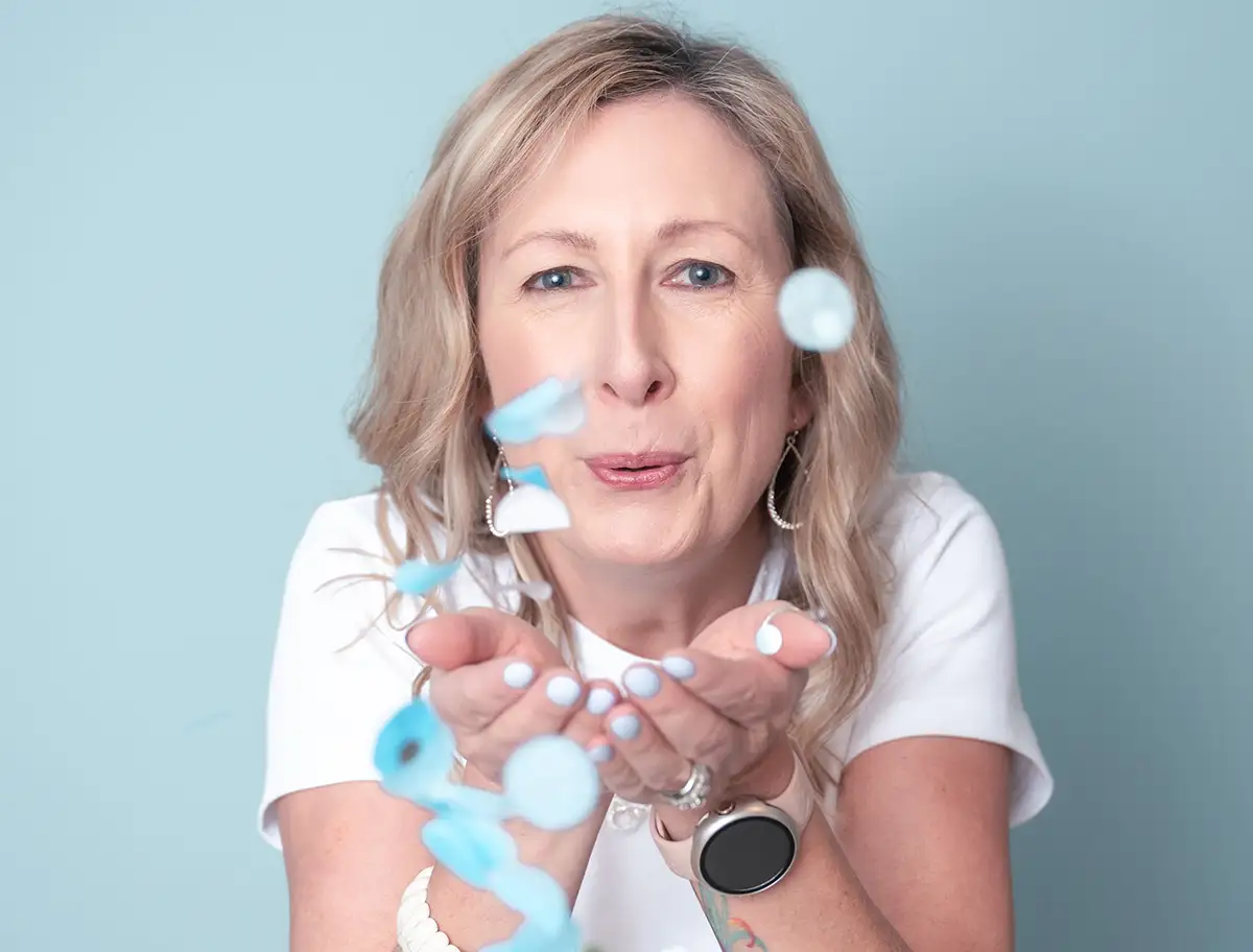 Erin Houston, a woman with blonde hair in a white shirt, blows blue confetti towards the camera against a light blue background. She wears hoop earrings, a smartwatch, and has light blue painted nails. - Favorite Grampy Travels