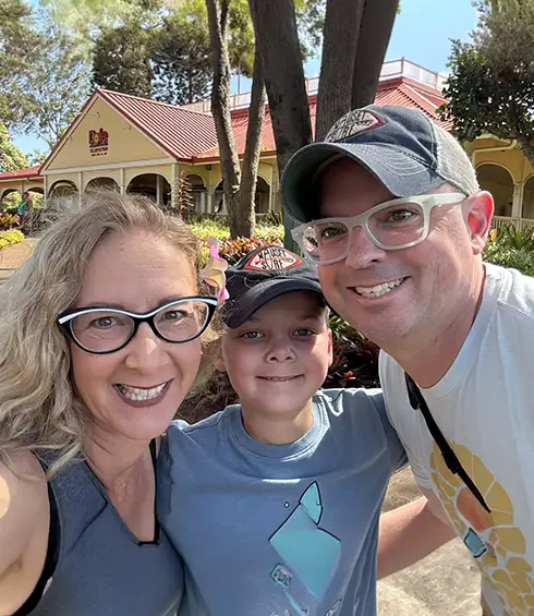 a Smiling Family of Three—two Adults and a Child—pose Together Outdoors in Front of a Yellow Building with a Red Roof and Trees Around Them on a Sunny Day. - Favorite Grampy Travels - Favorite Grampy Travels 2026