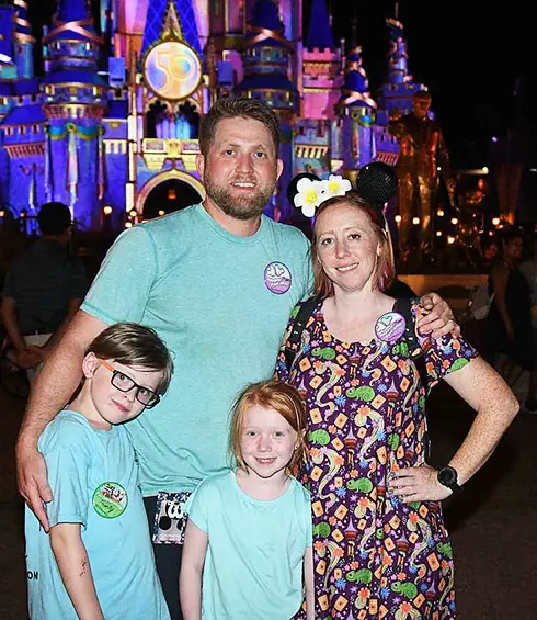 a Family of Four Poses in Front of a Brightly Lit Cinderella Castle at Night. the Adults Stand Behind Two Children, All Smiling. the Woman Wears Minnie Mouse Ears, and Everyone is Dressed Casually with Park Buttons on Their Shirts. - Favorite Grampy Travels - Favorite Grampy Travels 2026