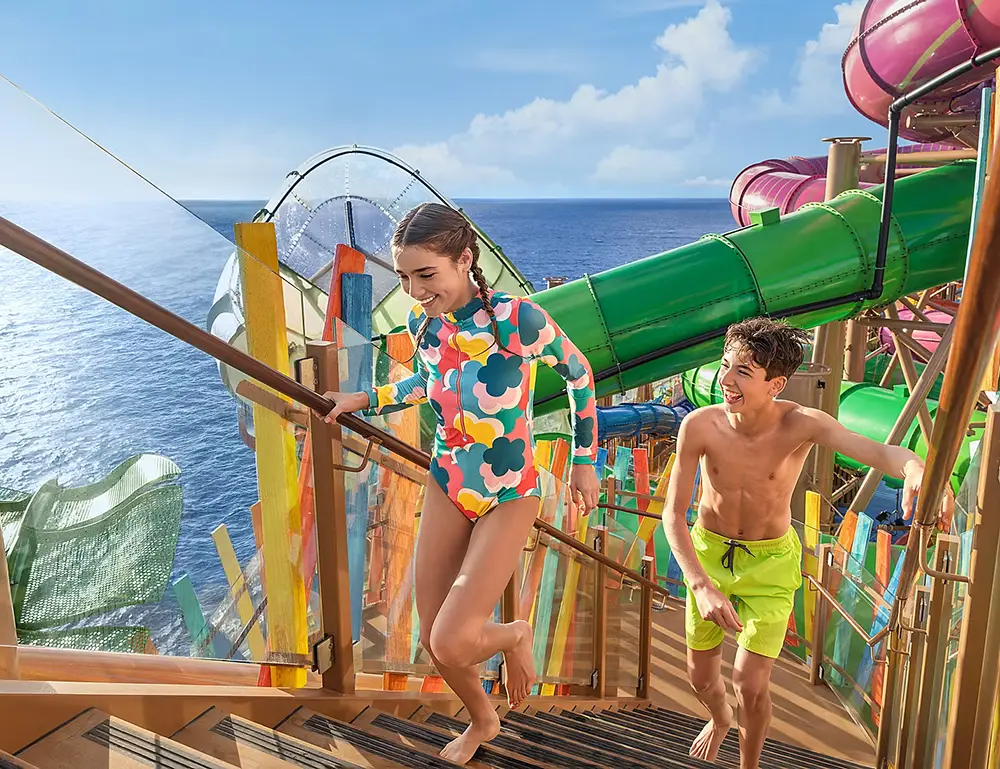 Two Kids in Swimwear Run Up Wooden Stairs Toward Water Slides on a Sunny Day Aboard the Star of the Seas, with Colorful Slide Tubes and the Ocean As Their Backdrop—perfect for a Family Cruise and the Ultimate Cruise Experience. - Favorite Grampy Travels