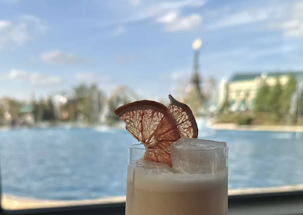 A close-up of a creamy drink with ice and dried citrus slices as garnish, set against a blurred background of a pool and distant buildings under a blue sky—a refreshing scene inspired by Celestial Park Dining. - Favorite Grampy Travels