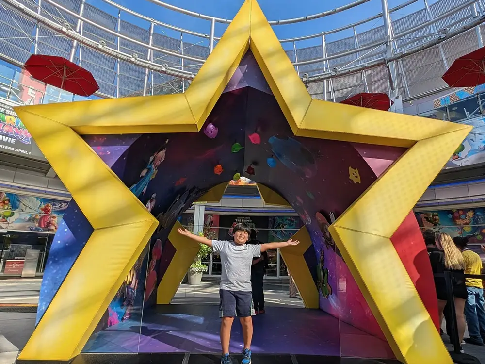 a Child Stands Smiling with Arms Outstretched Inside a Large, Yellow Star-shaped Structure at an Outdoor Venue, Evoking the Whimsy of a Family Movie Like Super Mario Galaxy Movie, with Colorful Decorations and People in the Background. - Favorite Grampy Travels