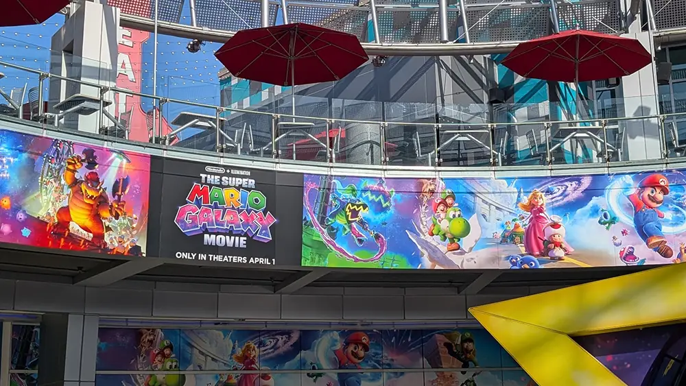 Curved Outdoor Billboard Displays Colorful Posters for the Super Mario Galaxy Movie, a Family Movie Featuring Mario, Bowser, Luigi, and Princess Peach in Vibrant Action Scenes. Red Umbrellas and Glass Railings Are Above. It’s Definitely Worth Seeing!. - Favorite Grampy Travels