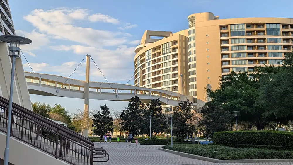 a Modern Curved Hotel, Disney's Contemporary Resort, with many balconies stands behind a skybridge walkway. People stroll along a tree-lined, paved path in the foreground under a blue sky—perfect for Disney World planning. - Favorite Grampy Travels