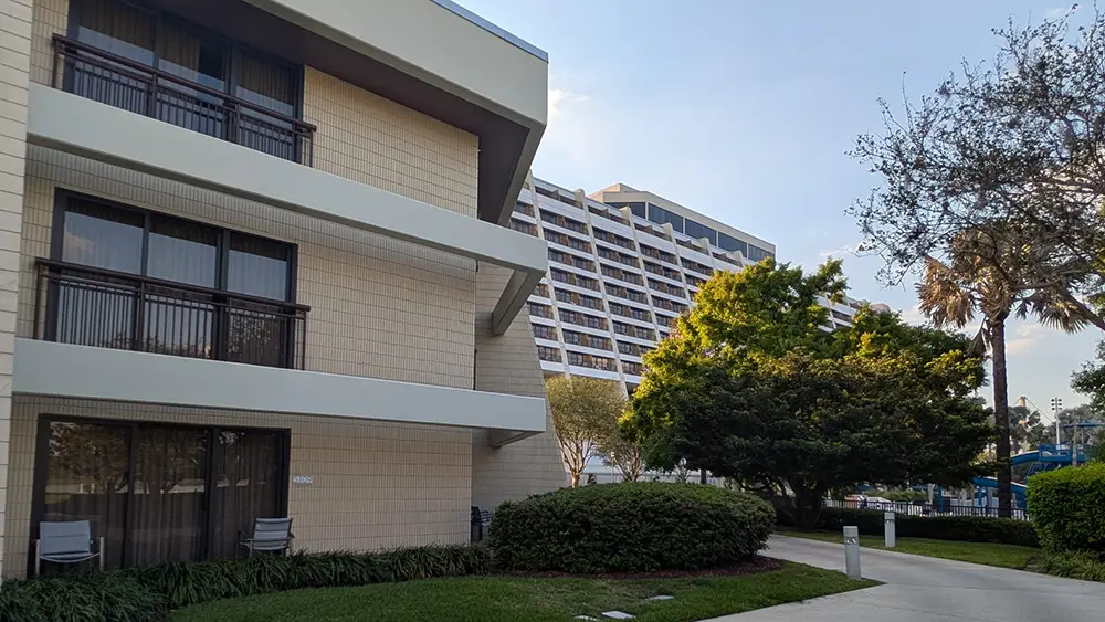 a Three-story Beige Building with Balconies Stands in the Foreground, While Disney's Contemporary Resort, a taller, modern, triangular hotel—one of the top Disney World Deluxe Resorts—rises in the background amid green trees and a walkway. - Favorite Grampy Travels