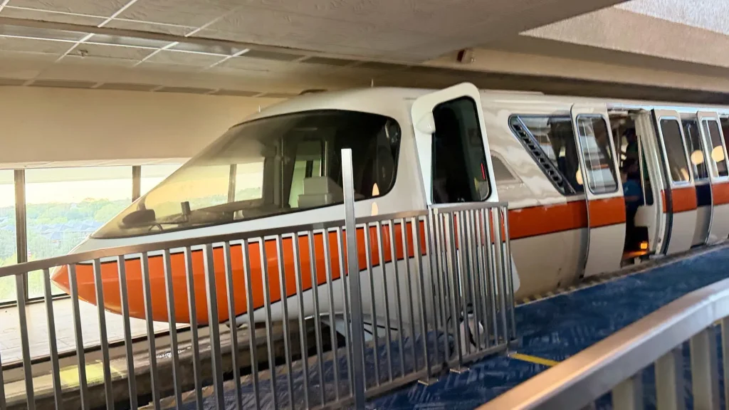 a White Monorail with an Orange Stripe is Stopped at an Indoor Station Platform at Disney's Contemporary Resort, with metal railings and blue carpeting visible in the foreground—a classic sight during Disney World planning or visits to Deluxe Resorts. - Favorite Grampy Travels