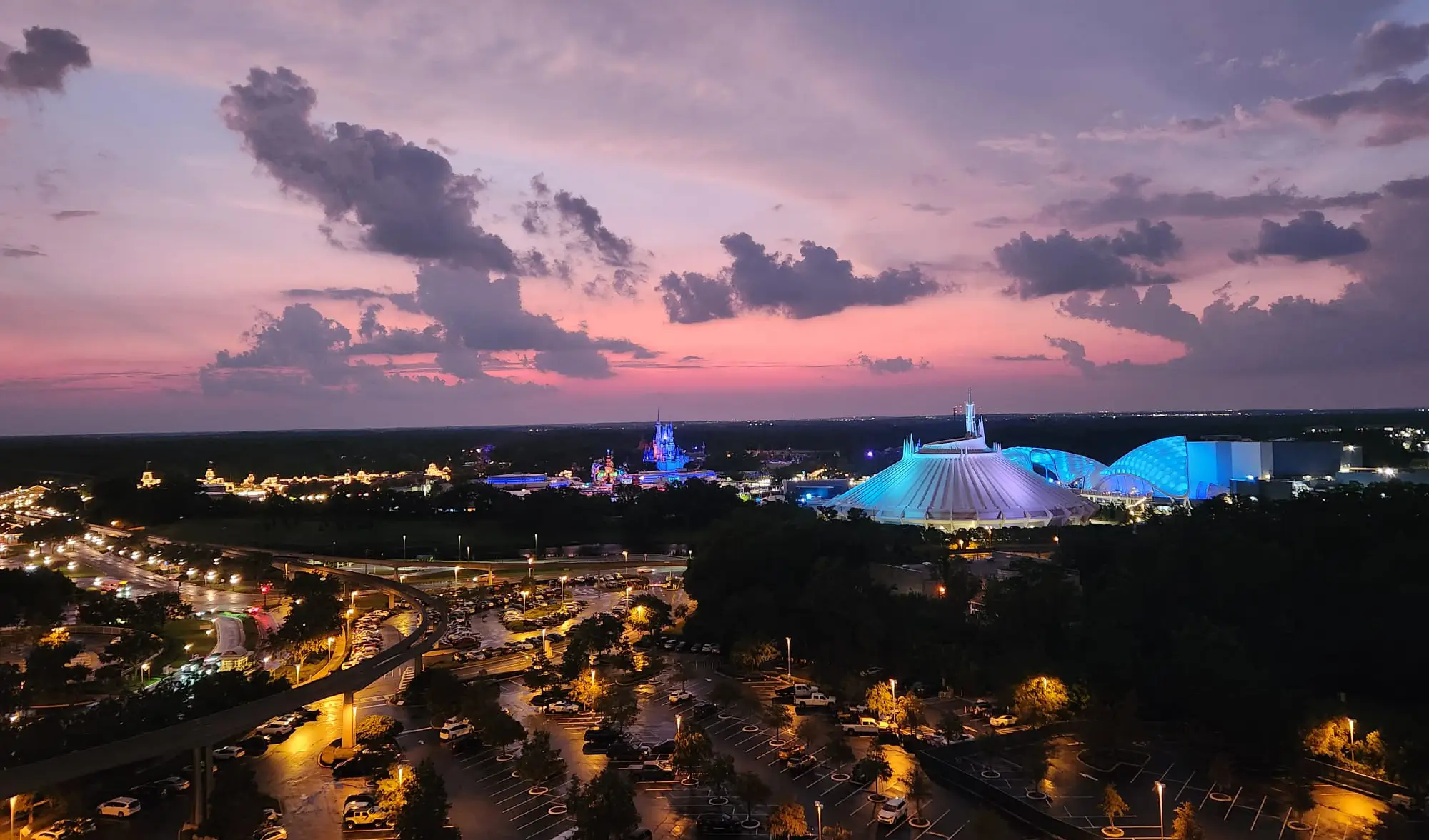 a View of a Theme Park at Sunset, Featuring Illuminated Buildings and Rides, Including a White Futuristic Dome. the Pink and Purple Sky Glows Above Bay Lake Tower, One of the Iconic Disney World Deluxe Resorts, As Parked Cars Fill the Lot Below. - Favorite Grampy Travels