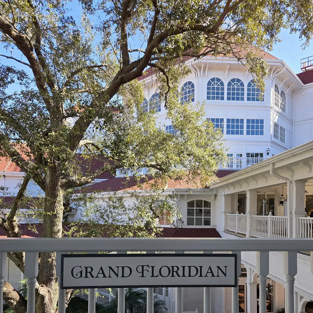 a View of the Grand Floridian Hotel’s White Victorian-style Building with Large Windows and Red Roof, Seen Through Leafy Trees and a Sign Reading “grand Floridian.”. - Favorite Grampy Travels - Favorite Grampy Travels 2026