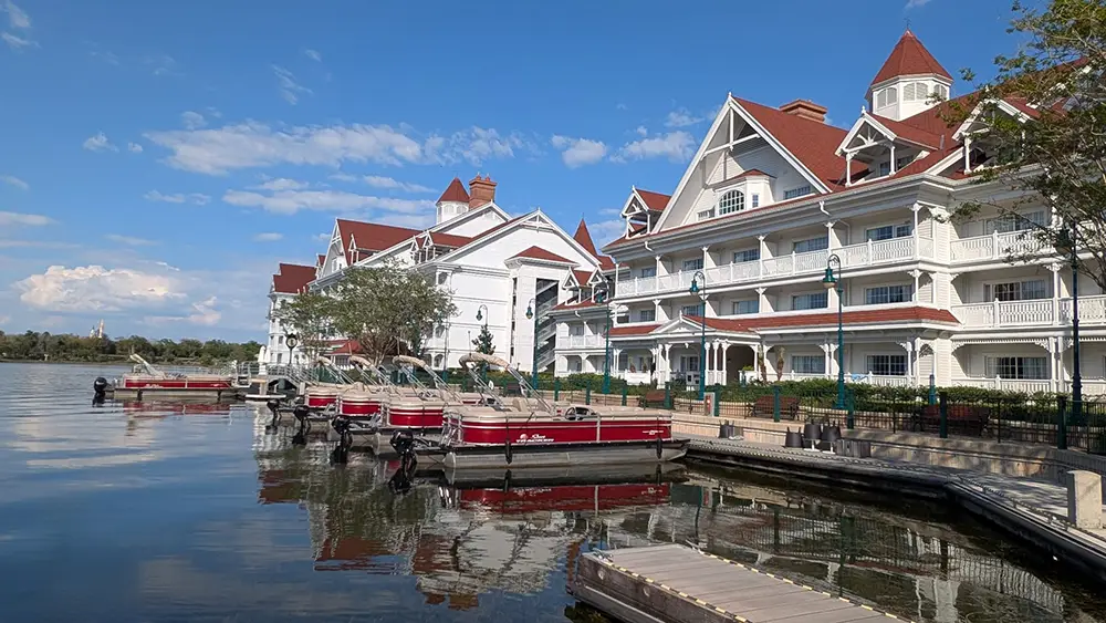 Disney's Grand Floridian Resort, a large white Victorian-style hotel with red roofs, sits beside a calm lake where red and white pontoon boats are docked. Perfect for a family guide to your next Disney resort getaway under blue skies and scattered clouds. - Favorite Grampy Travels