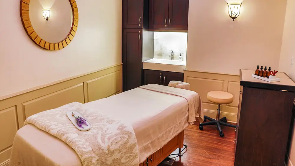 a Cozy Massage Room at Disney's Grand Floridian Resort features a neatly made table, wooden mirror, small sink area, cabinets, rolling stool, and bottles of essential oils. Warm lighting adds to the relaxing atmosphere—perfect for any family guide to unwind. - Favorite Grampy Travels