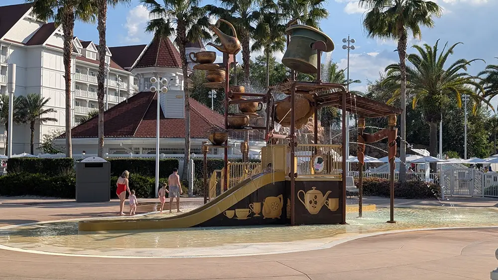 a Children’s Splash Pad at Disney's Grand Floridian Resort features a teapot-themed water play structure with slides and tipping buckets. Several kids and an adult are nearby, with palm trees and a large hotel building in the background. - Favorite Grampy Travels