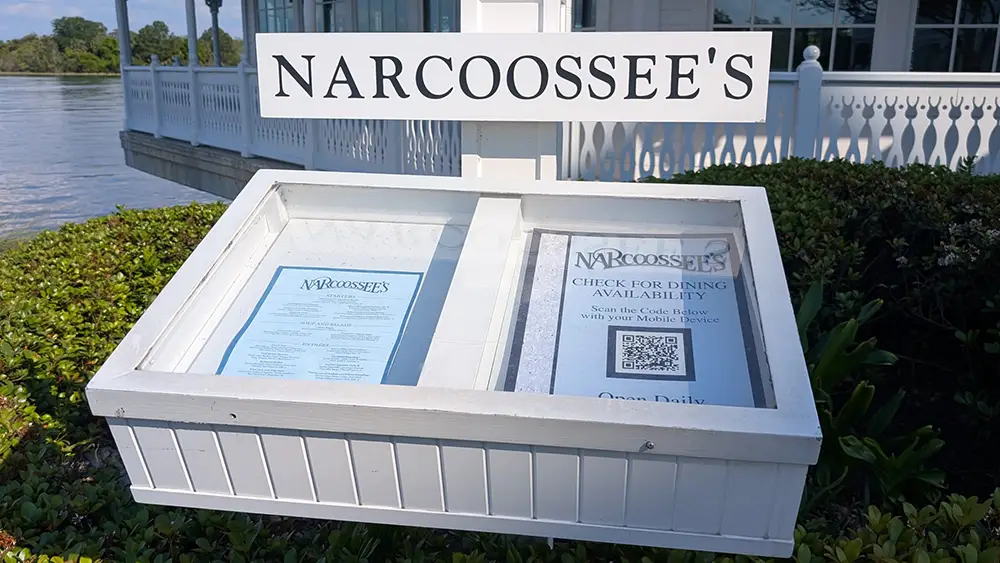 a White Outdoor Display Case at Disney's Grand Floridian Resort shows Narcoossee’s restaurant menus, one printed and one with a QR code for dining availability, in front of a waterfront building with white railings and greenery. - Favorite Grampy Travels