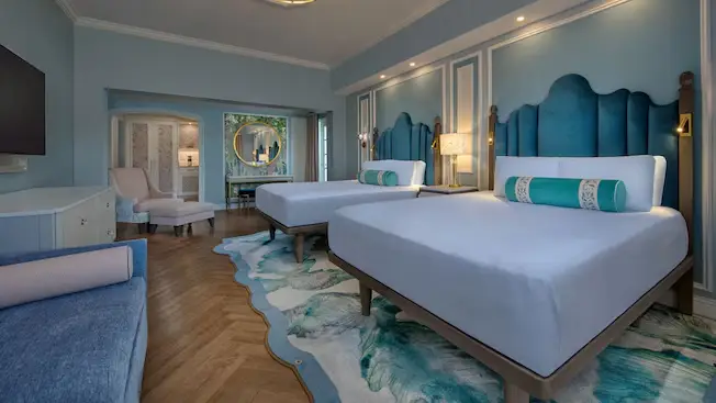 a Hotel Room at Disney's Grand Floridian Resort with two queen beds, blue upholstered headboards, aqua and white decor, a modern rug, a large wall mirror, lounge chair, wooden floors, and soft lighting—perfect for families. - Favorite Grampy Travels
