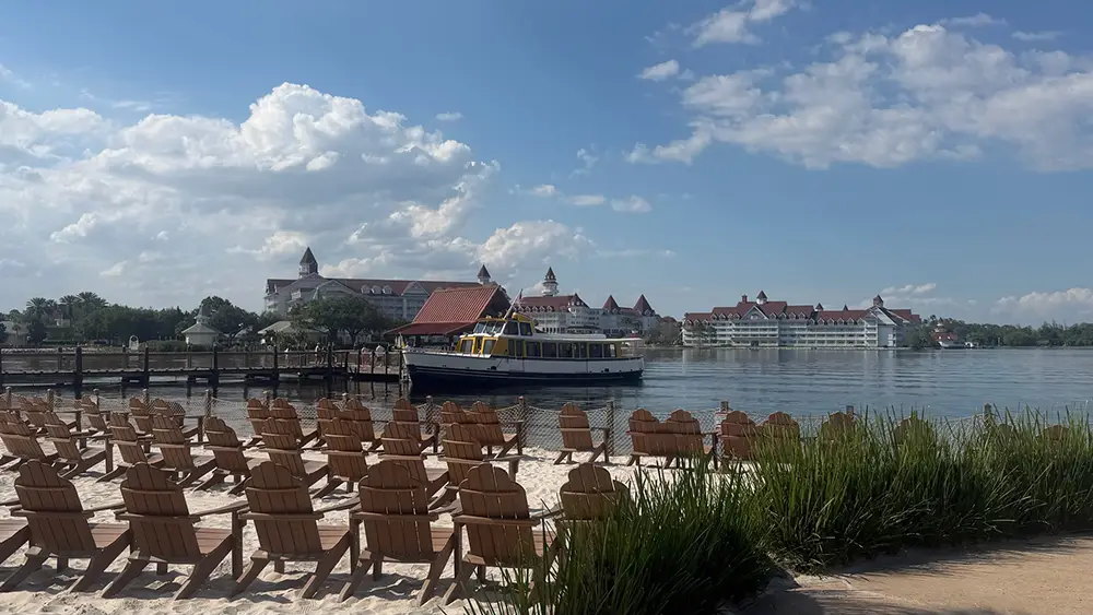 Rows of Empty Wooden Chairs Face a Calm Lake with a Boat on the Water; Large, Elegant Buildings with Red Roofs and Towers, Reminiscent of Disney's Polynesian Village Resort, stand in the background under a partly cloudy sky. - Favorite Grampy Travels