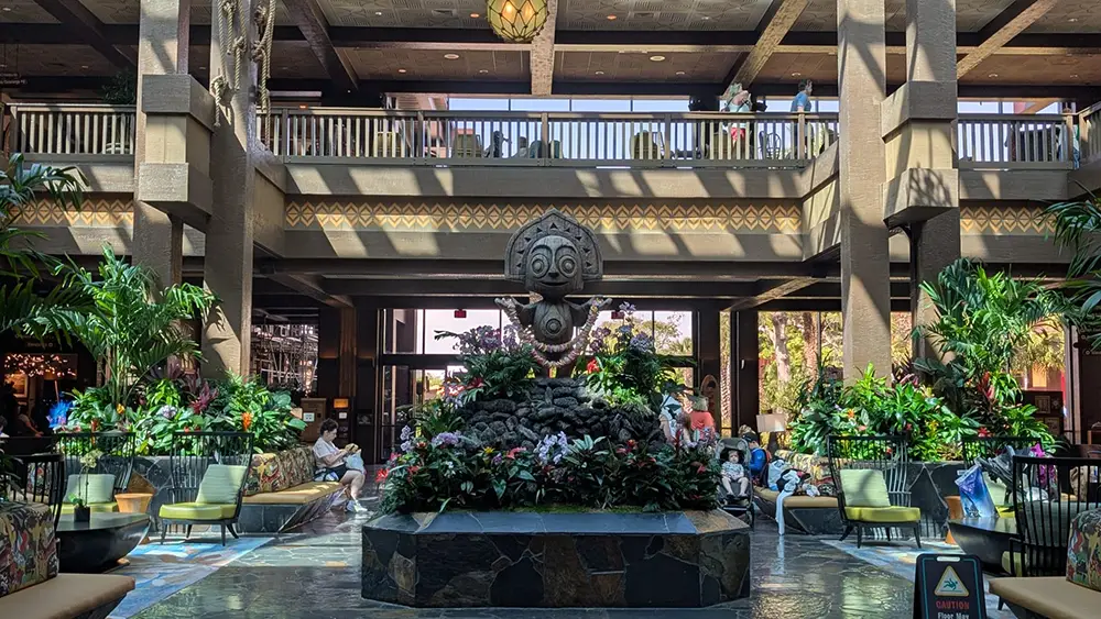 a Spacious Indoor Atrium at Disney's Polynesian Village Resort filled with tropical plants and a central stone fountain featuring a tiki statue, surrounded by seating areas and natural sunlight streaming from above. People are relaxing and walking in the background. - Favorite Grampy Travels