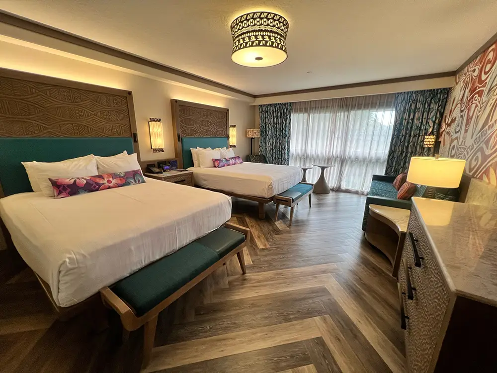 a Hotel Room at Disney's Polynesian Village Resort with two large beds, colorful accent pillows, wooden furniture, a patterned ceiling light, decorative wall art, a TV on a dresser, and large windows with curtains letting in natural light. - Favorite Grampy Travels