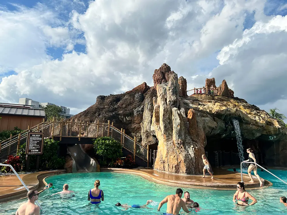 Guests Swim and Unwind in the Pool at Disney's Polynesian Village Resort, surrounded by a large artificial rocky volcano. A small waterfall cascades from the rocks, and a staircase winds up the side under a partly cloudy sky. - Favorite Grampy Travels