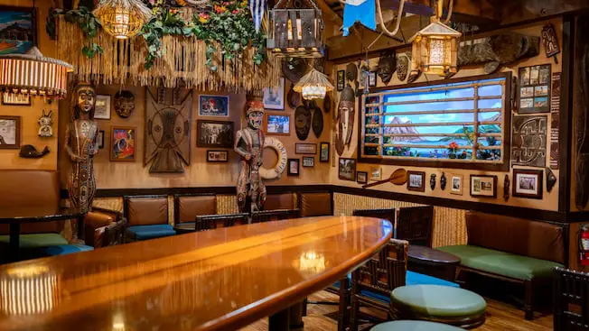 a Tiki Bar Decorated with Tropical and Polynesian-themed Decor, Wooden Statues, Framed Photos, Masks, Hanging Lanterns, Cushioned Seating, and a Mural of a Beach Scene Reminiscent of Disney's Polynesian Village Resort. - Favorite Grampy Travels