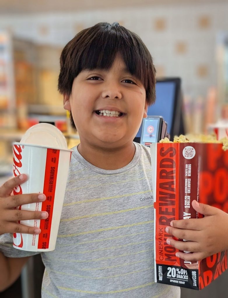 Smiling Boy in a Gray Shirt Holding a Large Soda Cup and a Box of Popcorn, Standing in What Appears to Be a Movie Theater Concession Area, Ready for a Family Outing to Watch the Worth Seeing Super Mario Galaxy Movie. - Favorite Grampy Travels