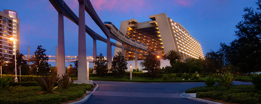 a Futuristic Hotel with a Brightly Lit Atrium and a Monorail Track Passing Through It, Surrounded by Greenery, Under a Purple and Blue Evening Sky—perfect for Disney World Planning at One of the Iconic Magic Kingdom Resorts. - Favorite Grampy Travels