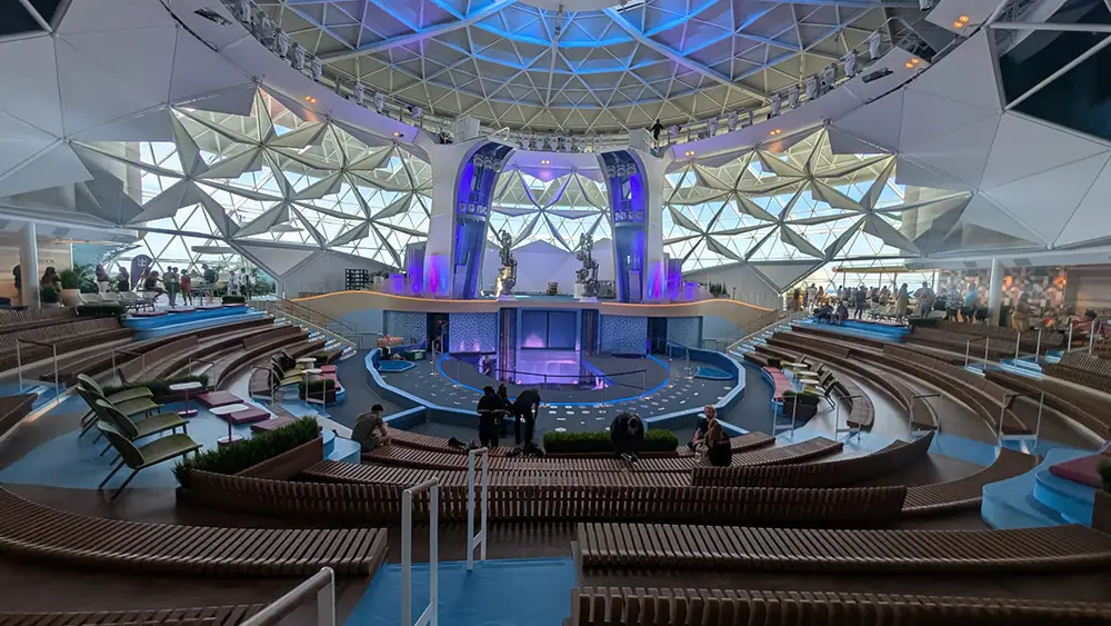 the Aquadome -modern Amphitheater with Tiered Seating, a Circular Stage, Geometric Glass Ceiling, and Purple-blue Lighting—perfect for Enjoying Entertainment on the Star of the Seas During Your Ultimate Cruise Experience. Decorative Plants and Lounge Chairs Line the Sides. - Favorite Grampy Travels