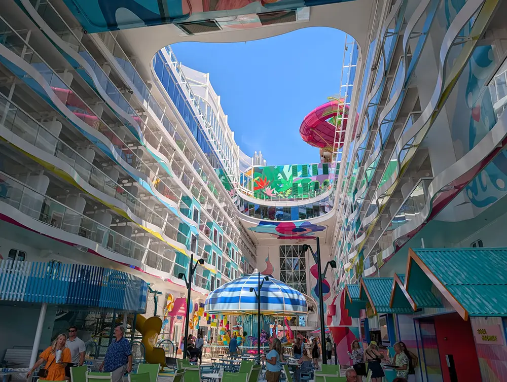 People Stroll and Relax in a Brightly Colored Open-air Area on the Star of the Seas Cruise Ship, Surrounded by Balconies, Vibrant Murals, Beach Umbrellas, and Decorative Buildings, with a Clear Blue Sky and Water Slides Above for the Perfect Family Cruise Experience. - Favorite Grampy Travels