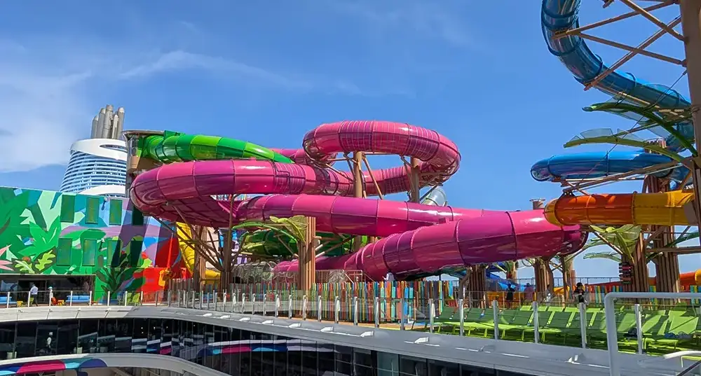 Colorful Water Slides, Mostly Pink and Green, Twist Around a Modern Outdoor Water Park Beneath a Blue Sky—perfect for a Family Cruise Adventure on Star of the Seas. a Tall, Contemporary Building Rises in the Background. - Favorite Grampy Travels
