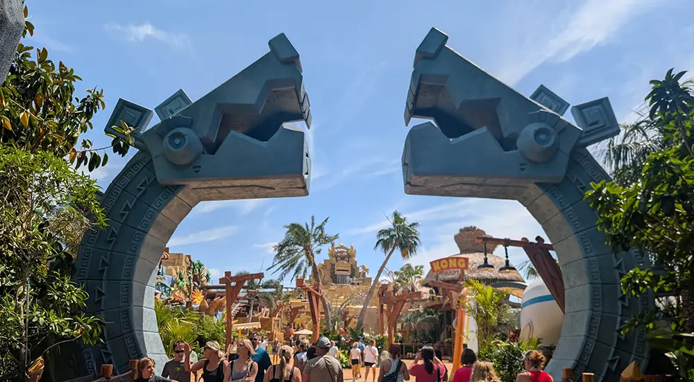 People Walk Under Two Large, Decorative Stone Archways Shaped Like Stylized Animal Heads in a Lively Outdoor Theme Park at Universal Orlando's Epic Universe, with palm trees and a bright blue sky. - Favorite Grampy Travels