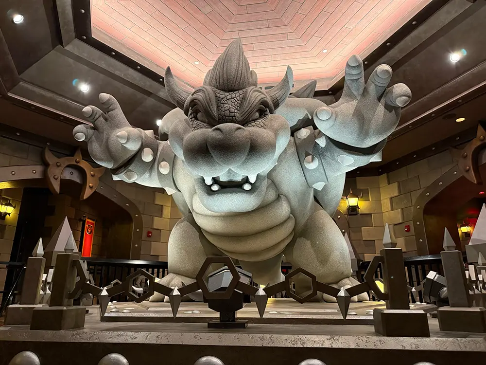 Giant Gray Stone Monster Statue with Horns and Spikes, Reaching Toward the Viewer from Behind a Spiked Barrier in a Dungeon-like Hall.