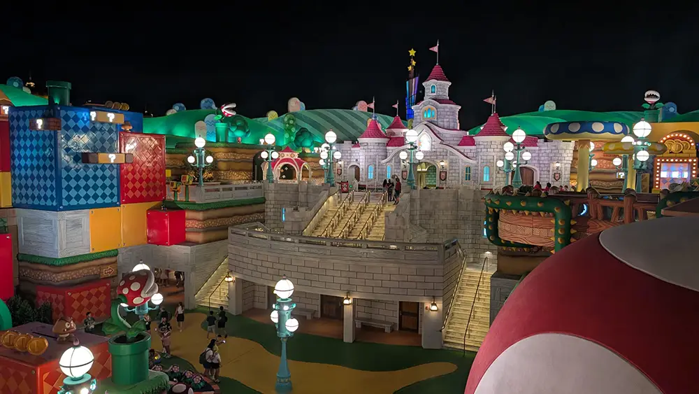 a Colorful, Whimsical Area at Night in Epic Universe’s Super Nintendo World, Featuring Blocky Buildings, Glowing Lamps, Large Mushrooms, and a Castle Reminiscent of Super Mario Games. Visitors Head Up Steps Toward Mario Kart Bowser's Challenge. - Favorite Grampy Travels