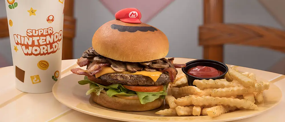 a Hamburger Topped with Bacon, Mushrooms, Cheese, Lettuce, and Tomato is Served with Crinkle-cut Fries, Ketchup, and a Super Nintendo World Cup—the Perfect Treat for Family Travel at Universal Orlando. the Burger Bun Features a Mario Hat Decoration on Top. - Favorite Grampy Travels