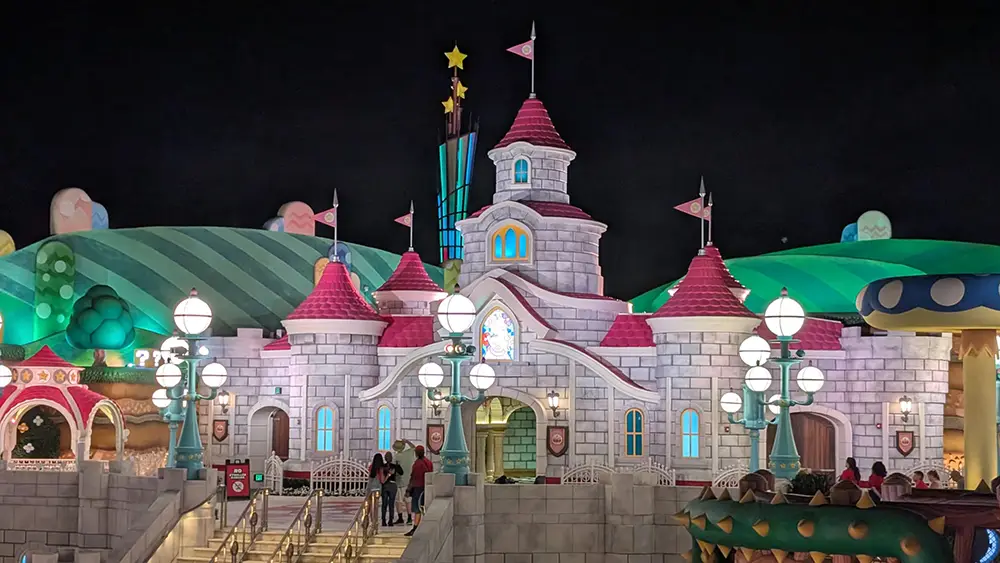 a Brightly Lit Fantasy Castle with Pink Roofs, White Walls, Flags, and Glowing Street Lamps Stands Against a Dark Sky, Surrounded by Whimsical Green Hills—a Magical Spot Worth Seeing for Any Family and Fans of the Super Mario Galaxy Movie. - Favorite Grampy Travels
