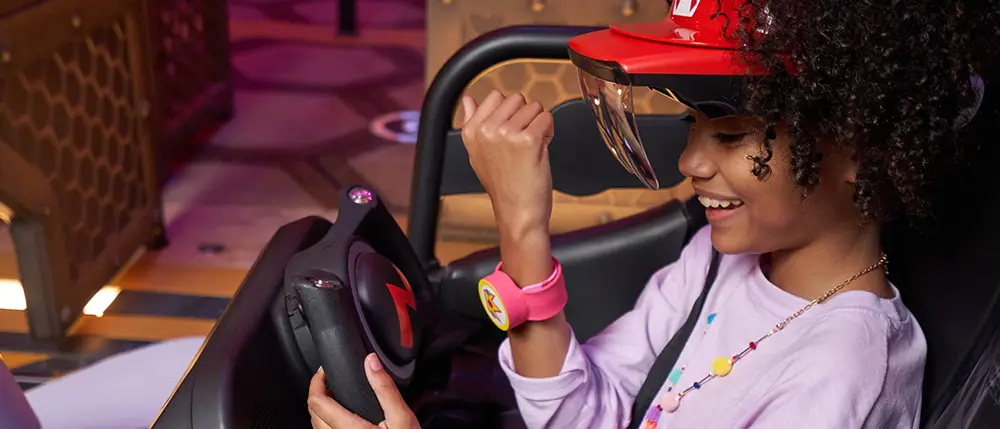 a Smiling Child Wearing a Red Visor and Pink Wristband Sits in a Theme Park Ride, Holding Onto the Steering Wheel and Raising One Arm with Excitement, Ready for Adventure on Mario Kart Bowser's Challenge. - Favorite Grampy Travels