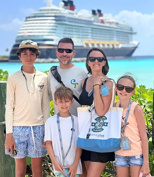 a Smiling Family of Five Poses Outdoors with a Disney Cruise Ship in the Background. the Parents and Three Children Wear Sunglasses and Casual Summer Clothes, Enjoying a Sunny Day Near the Beach at Disney's Castaway Cay, Disney Cruise Lines' Private Island - Favorite Grampy Travels - Favorite Grampy Travels 2026