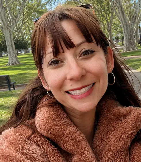 Nicole Damanio, a Travel Agent with Favorite Grampy Travels, is Pictured with Long Brown Hair and Bangs, Smiling at the Camera. She is Wearing a Brown Fuzzy Jacket and Hoop Earrings. Trees and Grass Fill the Background, Suggesting She is in a Park on a Sunny Day in Europe - Favorite Grampy Travels - Favorite Grampy Travels 2026