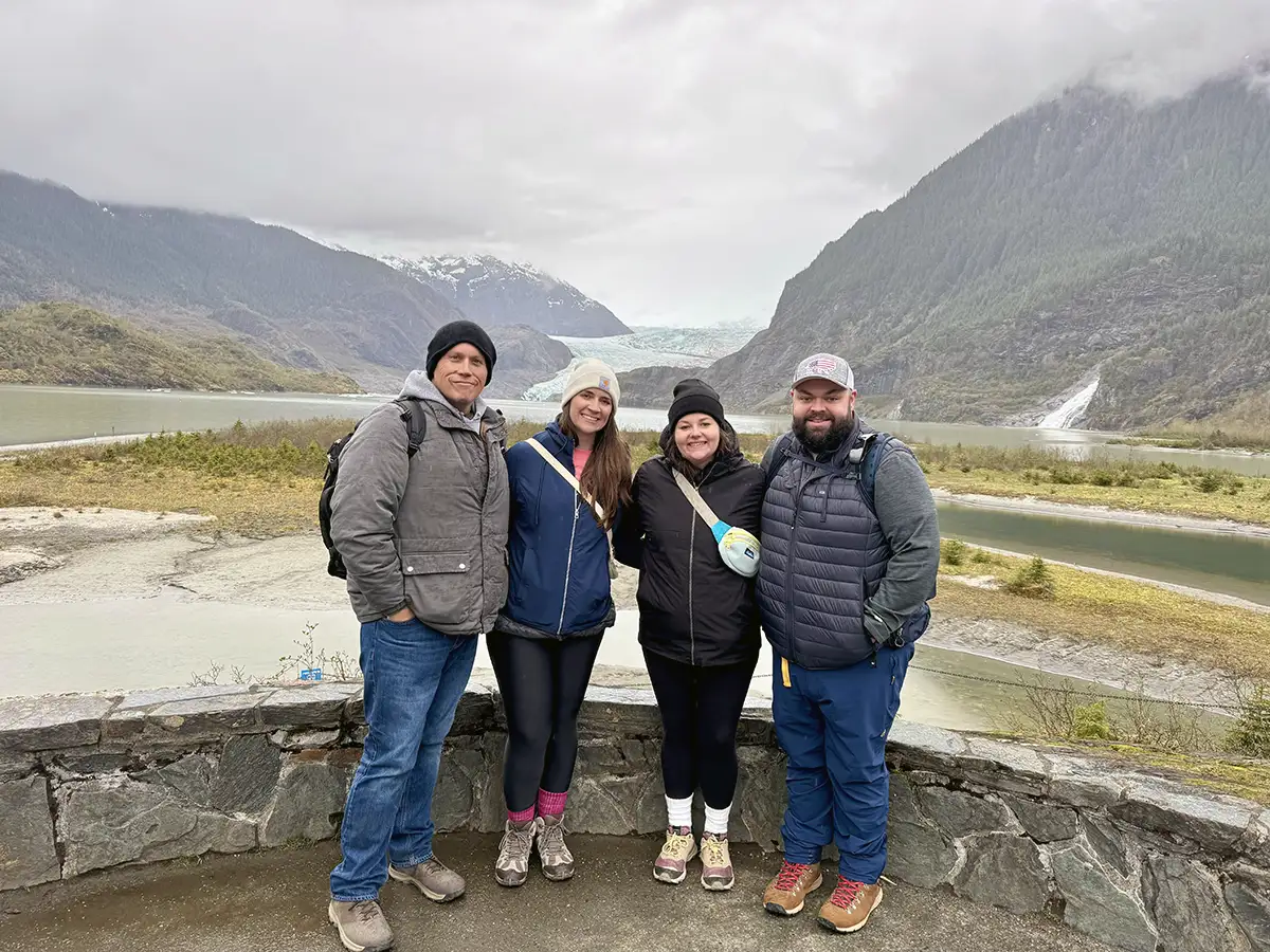 Samantha Malone and three family members, dressed in jackets and warm clothes, stand together smiling in front of a scenic backdrop of mountains, a glacier, and a winding river on a cloudy day. - Favorite Grampy Travels