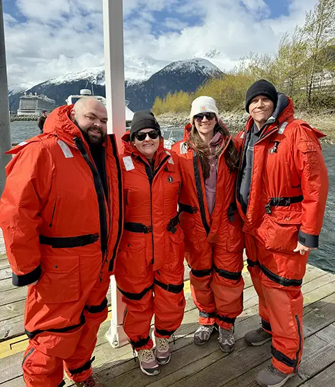 Samantha Malone and Her Family on Vacation Wearing Bright Orange Survival Suits Stand on a Dock by a River, with Snowy Mountains and an Anchored Cruise Ship in the Background Under a Partly Cloudy Sky. - Favorite Grampy Travels - Favorite Grampy Travels 2026