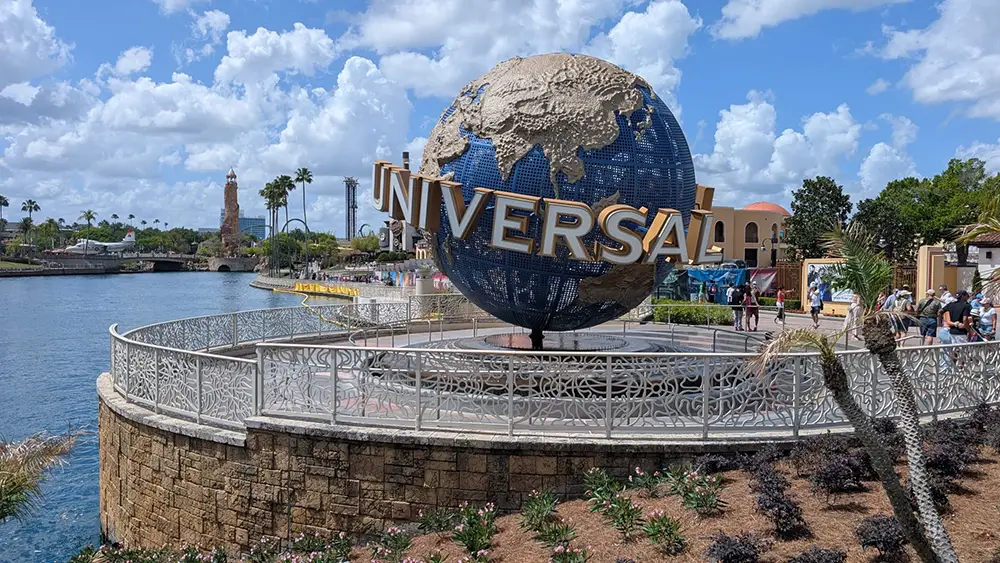 a Large Globe with the Word Universal Stands As a Landmark at the Entrance of Universal Orlando Resort, Surrounded by a Stone Planter, Water, Lush Landscaping, and Visitors Under a Partly Cloudy Sky. - Favorite Grampy Travels - Favorite Grampy Travels 2026