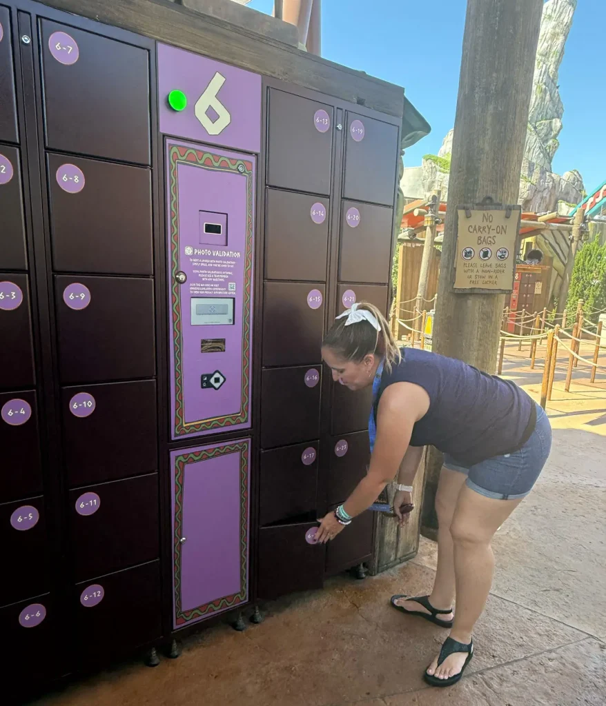 a Woman in Shorts and a T-shirt Bends Down to Open a Purple Universal Orlando Locker at an Outdoor Locker Station in an Amusement Park, with a Sign in the Background and a Ride Visible Behind Her. - Favorite Grampy Travels - Favorite Grampy Travels 2026