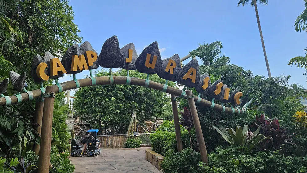 a Large Archway Reading Camp Jurassic with Rocky Decorations Stands Amid Lush Greenery and Tropical Plants at the Entrance of Universal Orlando’s Islands of Adventure. Two People with Strollers Are Visible Beneath the Arch. - Favorite Grampy Travels - Favorite Grampy Travels 2026