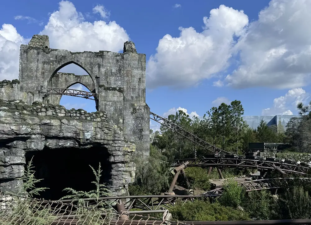 a Roller Coaster Track Winds Through Rocky Ruins and Lush Greenery at Universal Orlando Under a Bright Blue Sky. the Stone Ruins Resemble a Castle or Fortress, Partially Crumbled with an Arched Opening—just Like Hagrid - Favorite Grampy Travels 2026's Magical Creature Motorbike Adventure. - Favorite Grampy Travels
